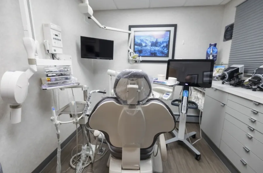 A bright treatment room with a pale grey chair and two monitors.